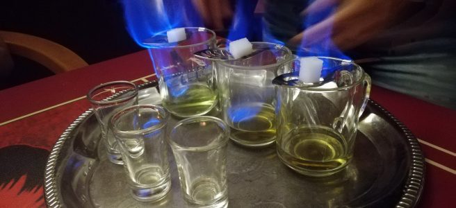 Three glasses with spoons balenced on them. both the green liquid and the sugar cubes on the spoon are on fire. the absinthe burns blue.