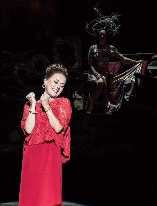 Tracie Bennett as Carlotta Campion. Photo by Johan Persson. In the foreground is the old Carlotta under a bright light in a tasteful red dress. In the background in dim lighting young Carlotta sits gracefully in a silver dress and stared crown. 