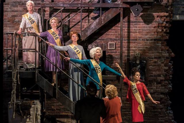 Josephine Barstow, Gemma Page, Janie Dee, Geraldine Fitzgerald and Tracie Bennett. Photo by Johan Persson. Five old women decend on a metal fire escape staircase attached to a crumbling brick wall. They are mostly happy. Each with a sash with the year they performed. 1918,1930,196,1938,1941. In the foreground is another old woman and her husband.