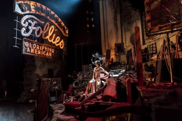 Emily Langham as Young Carlotta. Photo by Johan Persson. The dancer from 1938 sits on a bank of rubble and broken red furnishings. In the background a sign. Light bulbs, some burst, most burning bright, trace the words "Weismanns's Follies glorifying the american girl." The woman is dressed in silver and glitter. Her gown long and draping. Around her head are three rings with large six pointed stars on them.