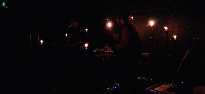 Dark stage with arc of nine filament light bulbs. Gif shows different lights glowing in rhythm, at times illuminating the two performers.