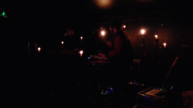 Dark stage with arc of nine filament light bulbs. Gif shows different lights glowing in rhythm, at times illuminating the two performers. 