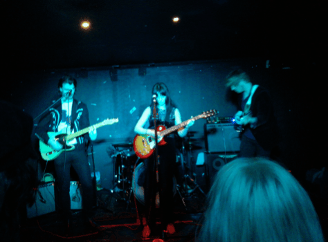 Fever machine in blue lighting. A woman head obscures party of the image. The three visible members stand on stage, all use guitars, there is a drummer but you can't see him