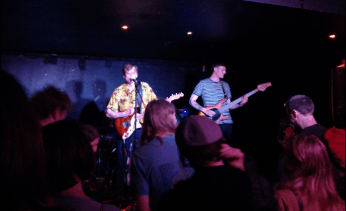 Gif of gladboy performing. People are swaying in the foreground. The lead singer is wearing a yellow and Hawaiian shirt, bassist is to his left in striped t-shirt and behind them a mop haired drummer in shirt and tie