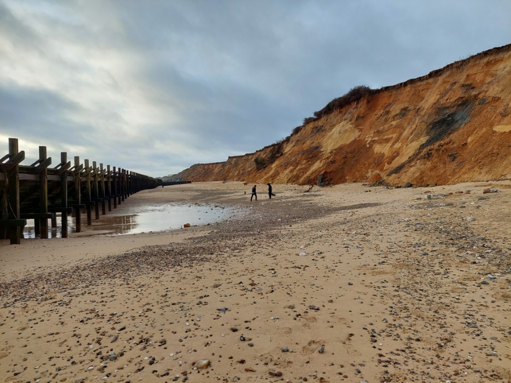 between wooden sea defences and crumbling pale brown cliffs is a sand beach with pebbles, a tide pool, and two men all in black in the distance. clumps of brambles have fallen part way down the cliffs.