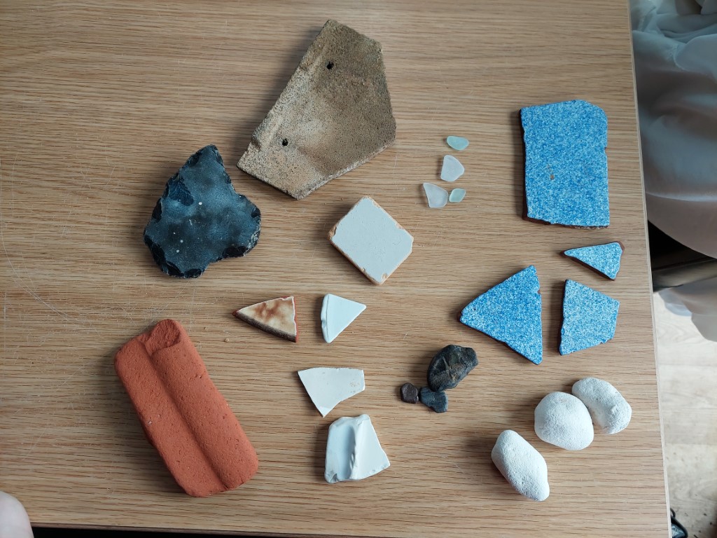 wooden table with fragments of white plates, blue speckled tile, terracotta plant pot, roof tile, white seaglass, and chalk lumps